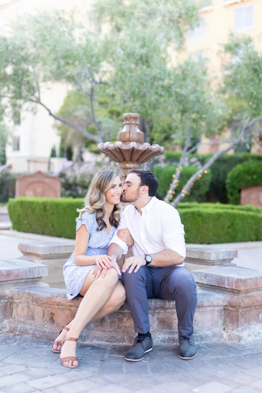Newly engaged couple laughing together on a stone bench by a fountain