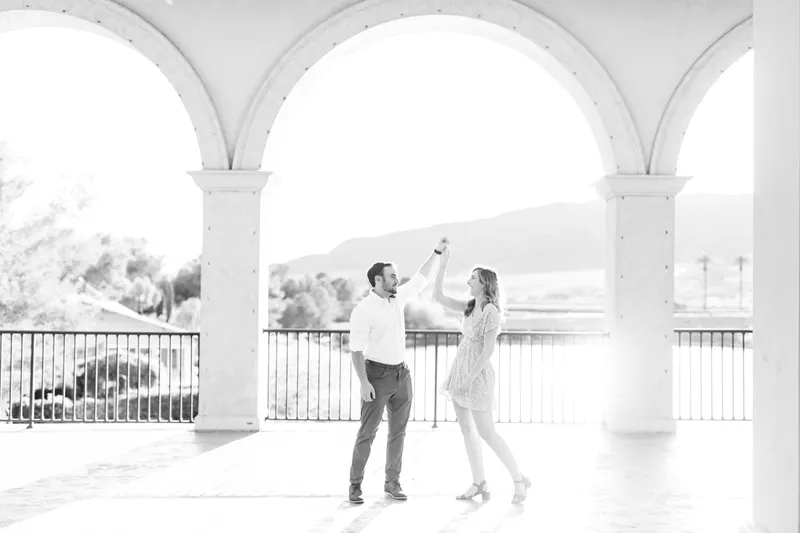 Black and white photo of a man spinning his fiancee under a row of tall stone arches above the water