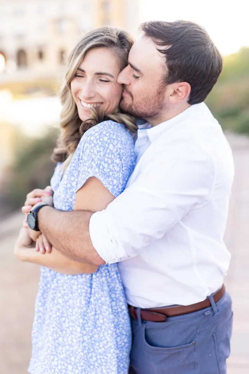 Couple holding each other in the late sun, him kissing the top of her head while she laughs with her eyes closed
