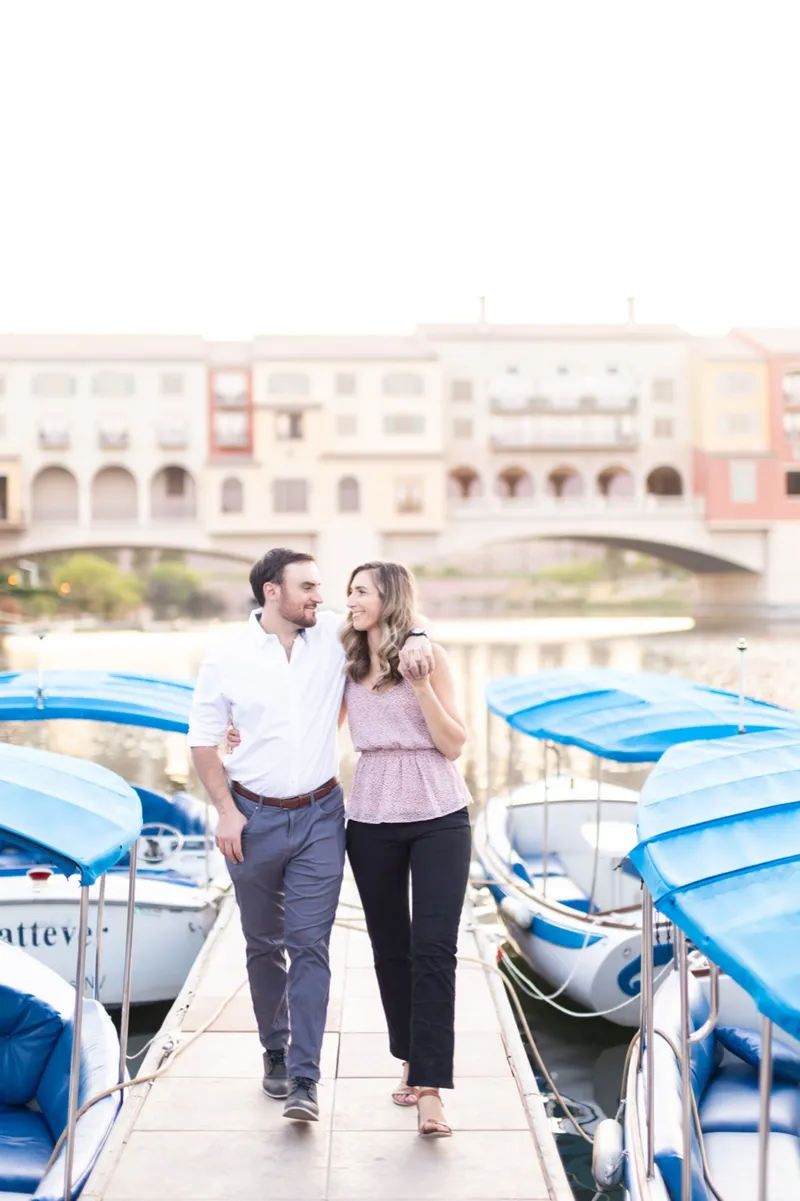 Couple walking a narrow dock between rows of boats under blue canopies with the village behind them