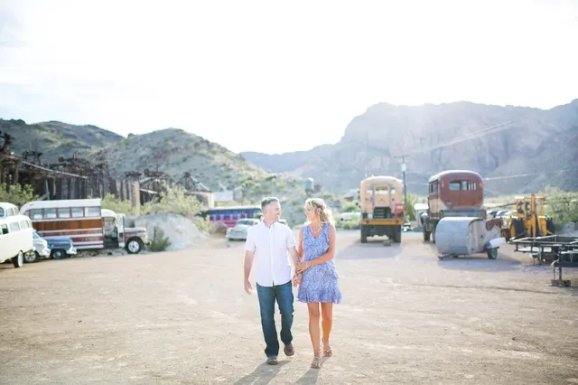 Couple walking hand in hand down a wide dirt lot lined with old buses and trucks, with a mining structure on the ridge above them