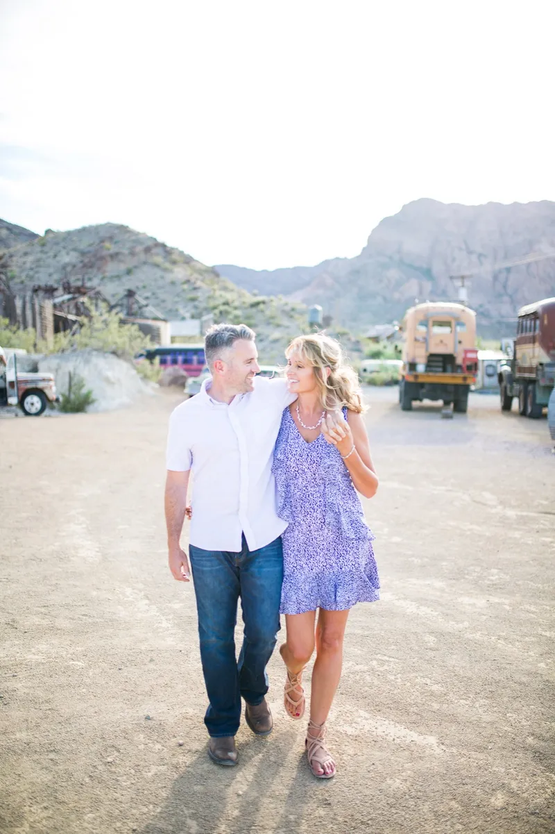 Couple walking arm in arm toward the camera across the gravel lot, both laughing, with parked trucks and desert hills behind them
