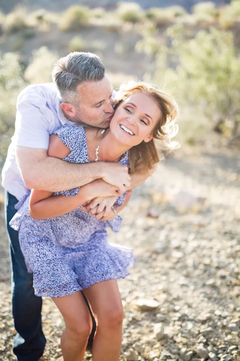 Man hugging his fiancee from behind in the desert brush and kissing her cheek while she laughs and her feet leave the ground