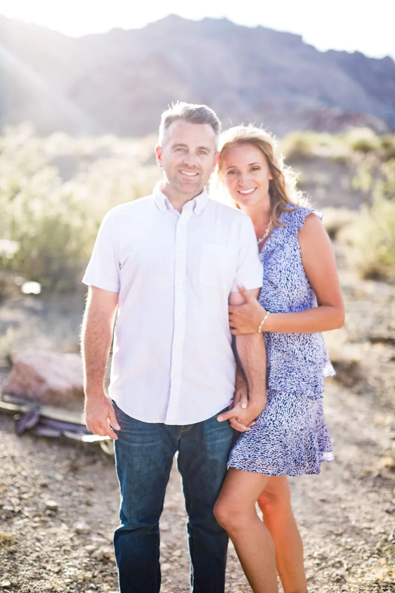 Couple standing arm in arm in the desert brush with dry hills behind them and the sun low over the ridge
