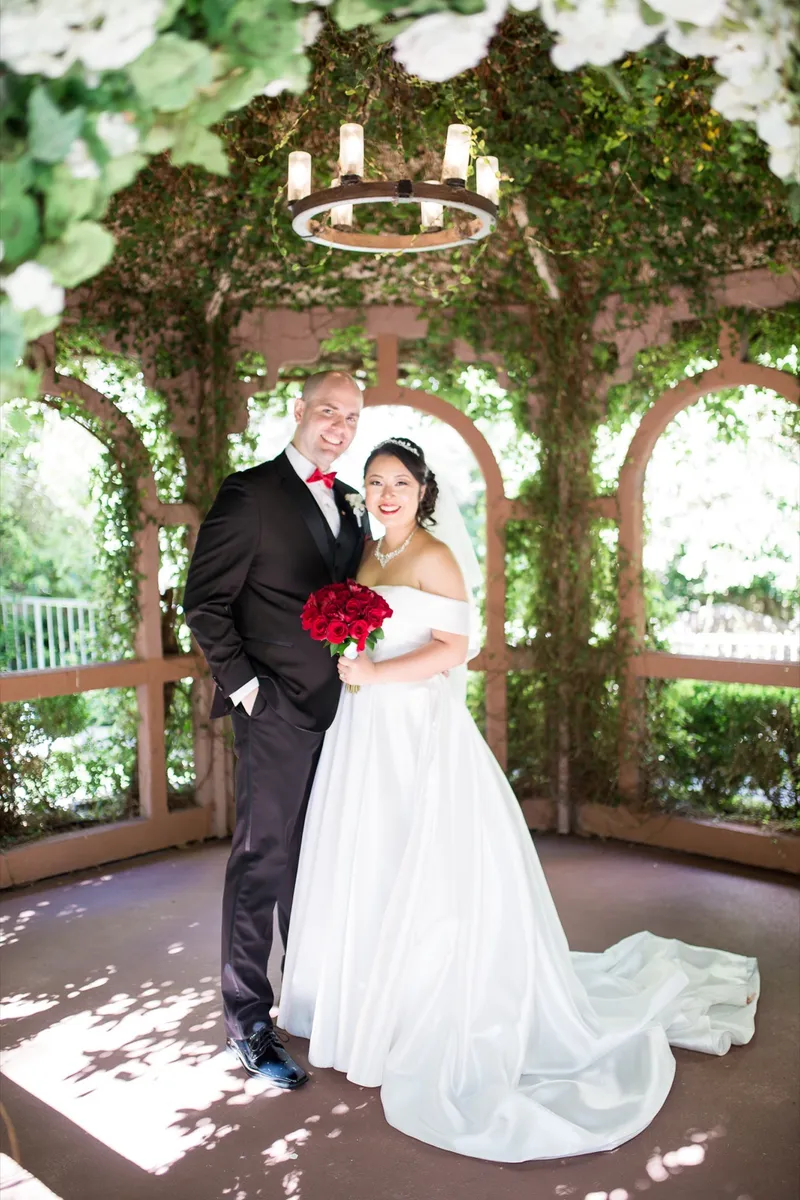Bride and groom standing together under the gazebo's iron chandelier, her red rose bouquet between them
