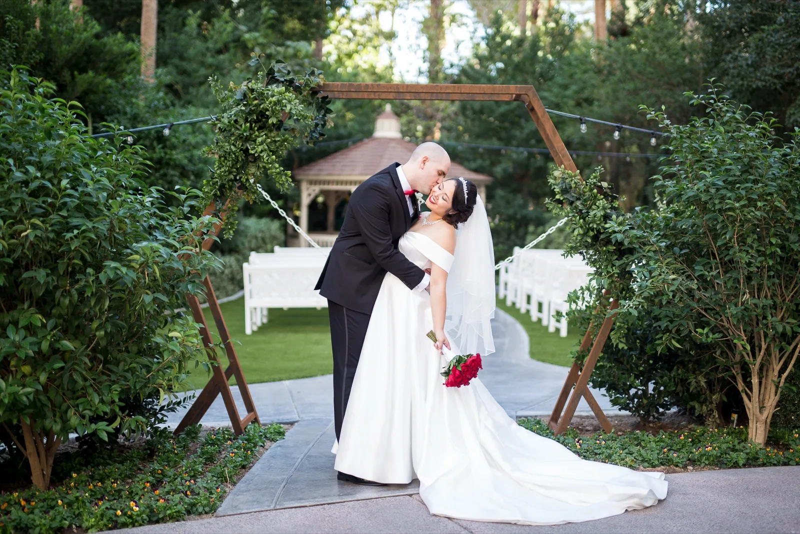 Groom dipping the bride into a kiss on the garden path with the ivy covered gazebo and rows of white chairs behind them