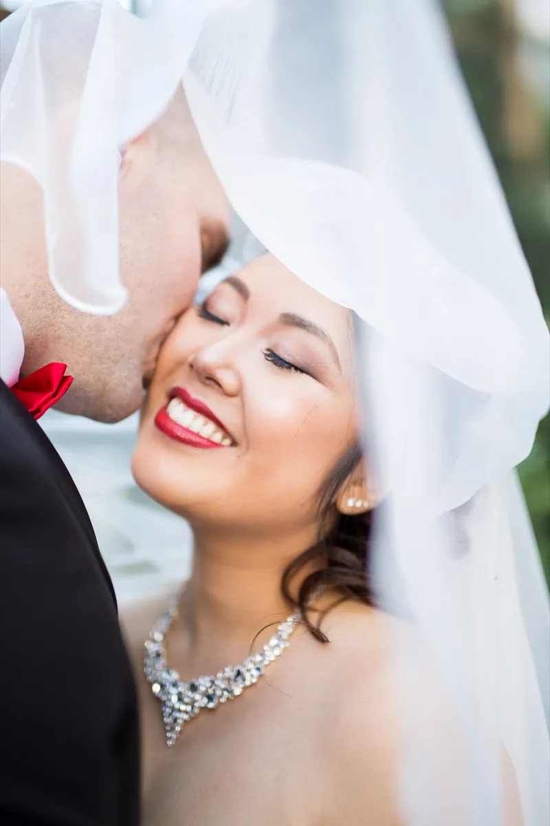 Close portrait of the bride laughing under her veil as the groom kisses her cheek