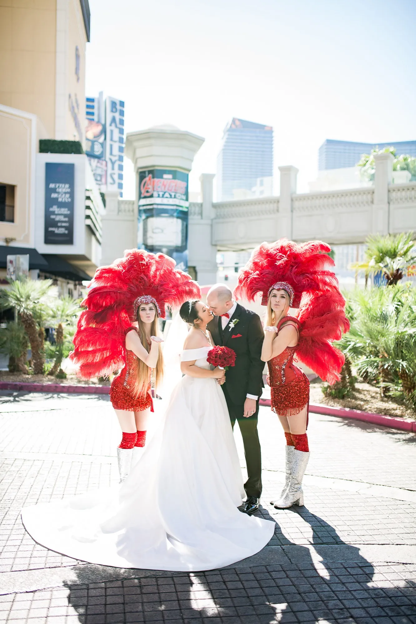 A bride and groom kissing on the Las Vegas Strip flanked by two showgirls in red feather headdresses