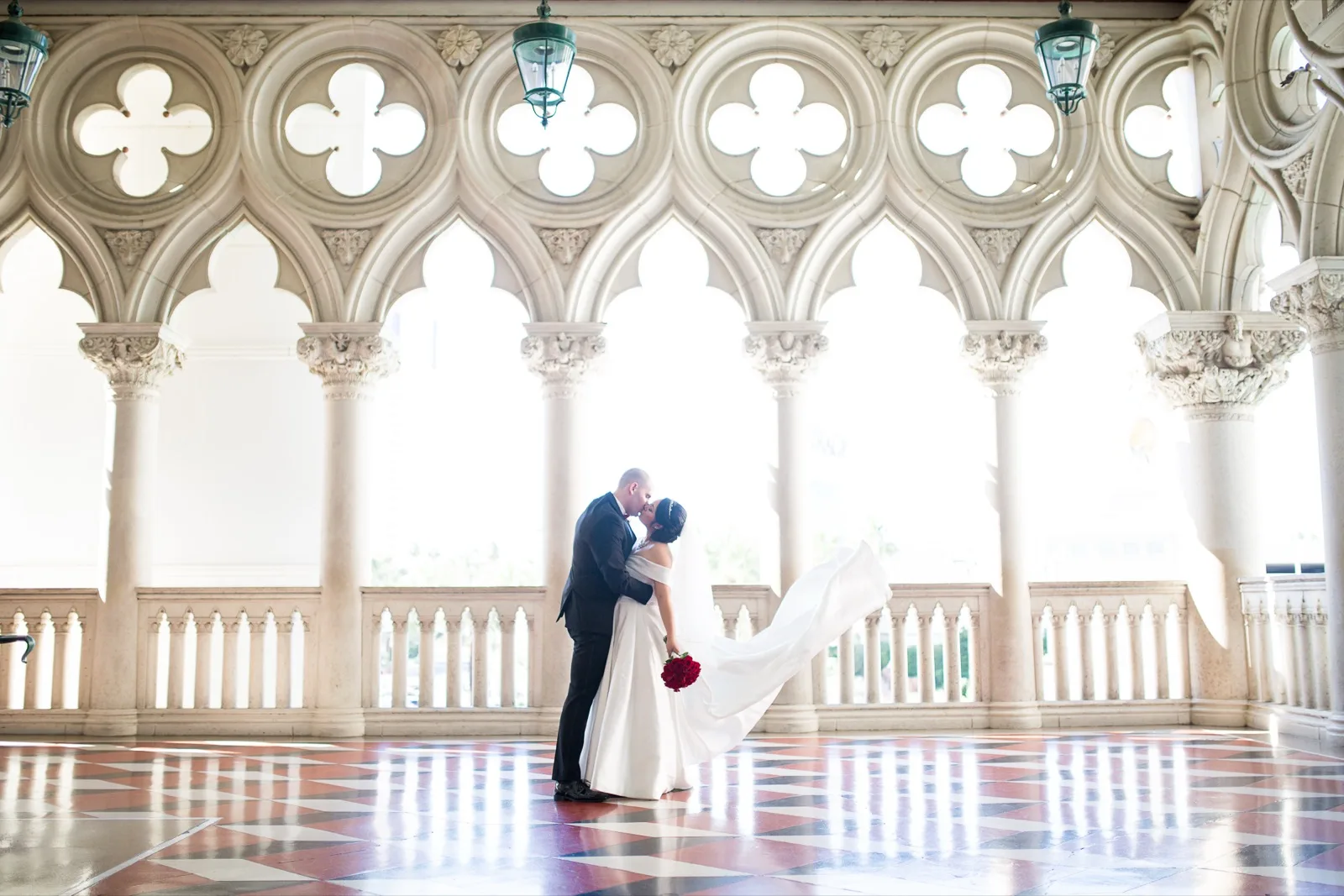 Groom dipping the bride into a kiss on the red and white checkered marble of the arched walkway, her train swinging out behind her