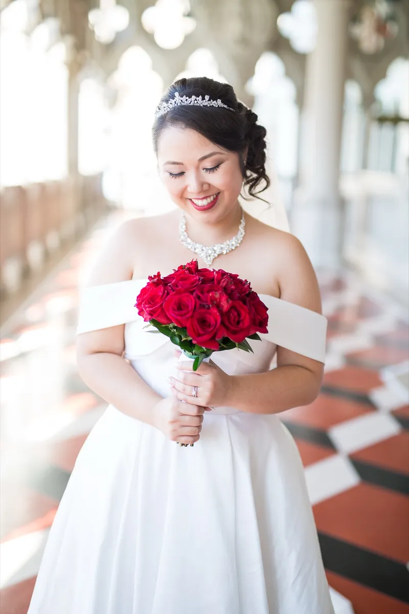 Bride in a tiara and off shoulder gown looking down at the bouquet of red roses in her hands