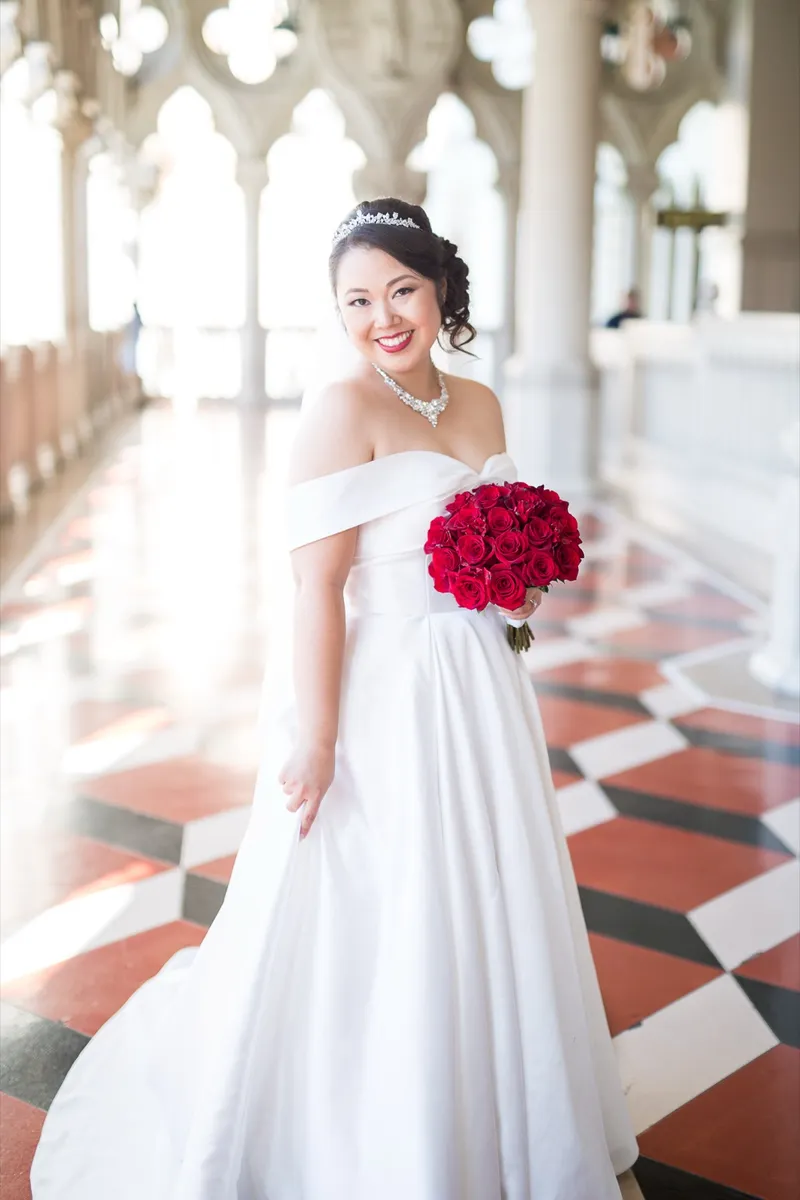 Bride smiling toward the camera with her red rose bouquet, pale columns and arches receding behind her