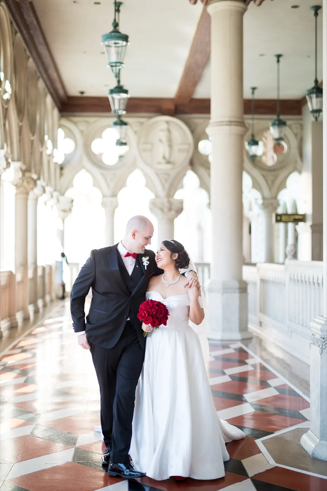 Bride and groom side by side in the arched walkway, his hand in his pocket, her red rose bouquet at her waist