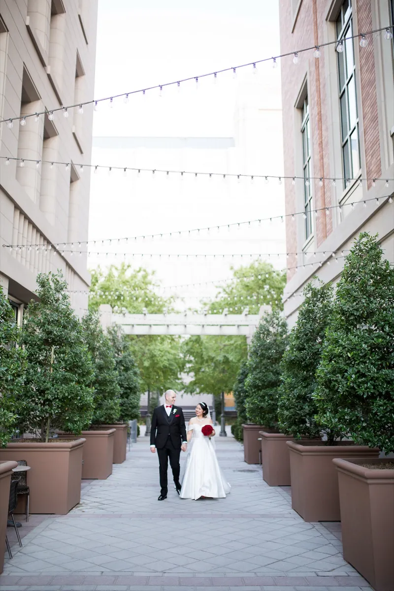 Bride and groom walking away down the string lit walkway, her red rose bouquet held at her side