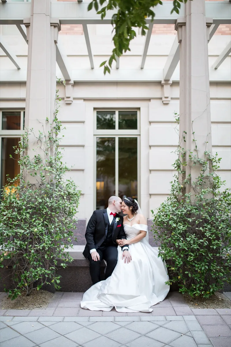 Bride in a satin ballgown sitting on the ledge beside her groom in the last of the evening light, green shrubs on either side