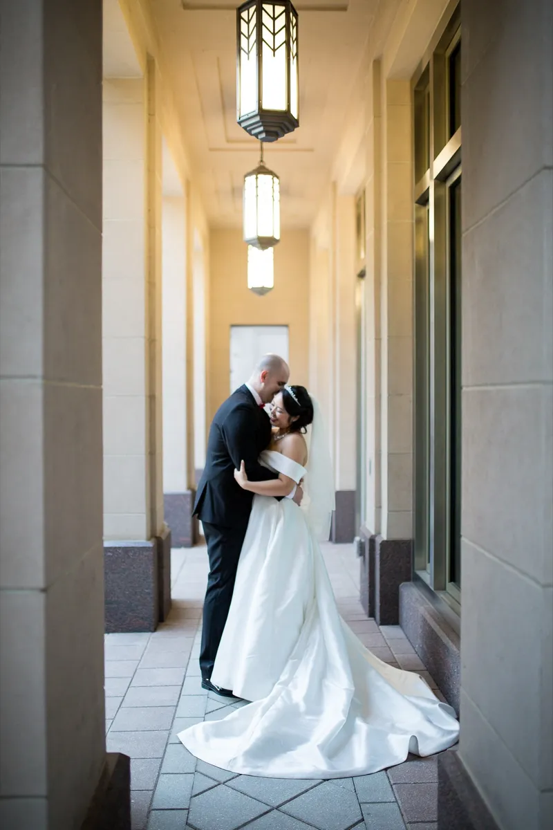 Groom leaning in to kiss his bride in the colonnade, her satin train pooling on the patterned tile behind them