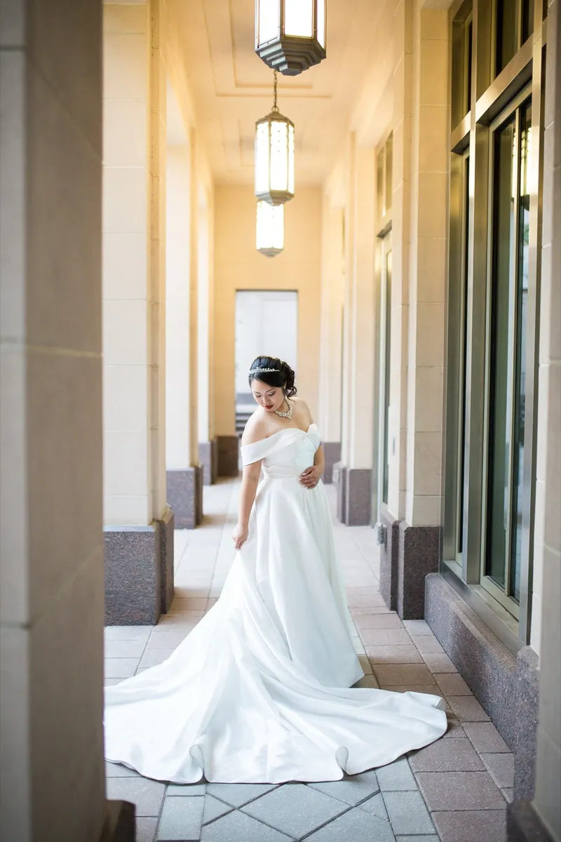 Bride holding out the skirt of her off the shoulder satin gown in a stone colonnade lit by hanging art deco lanterns
