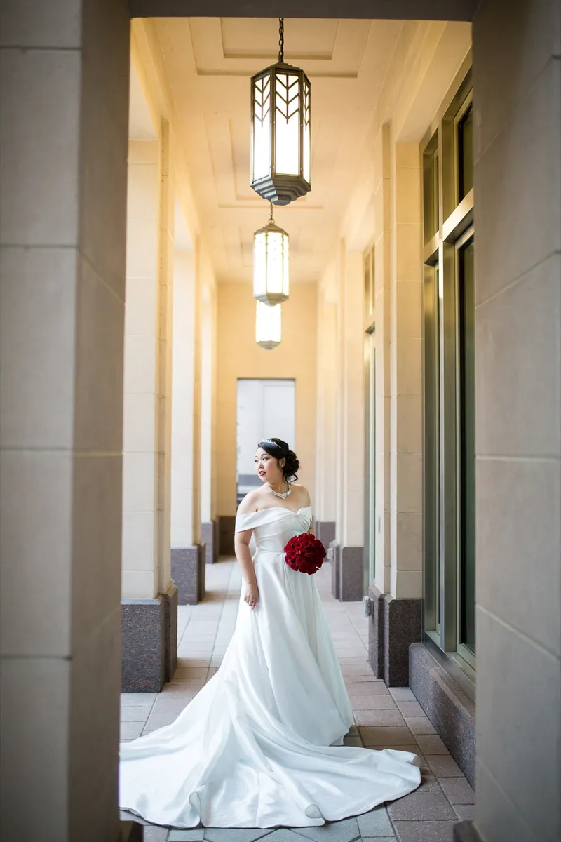 Bride standing between the stone columns holding a bouquet of red roses, lanterns glowing above her