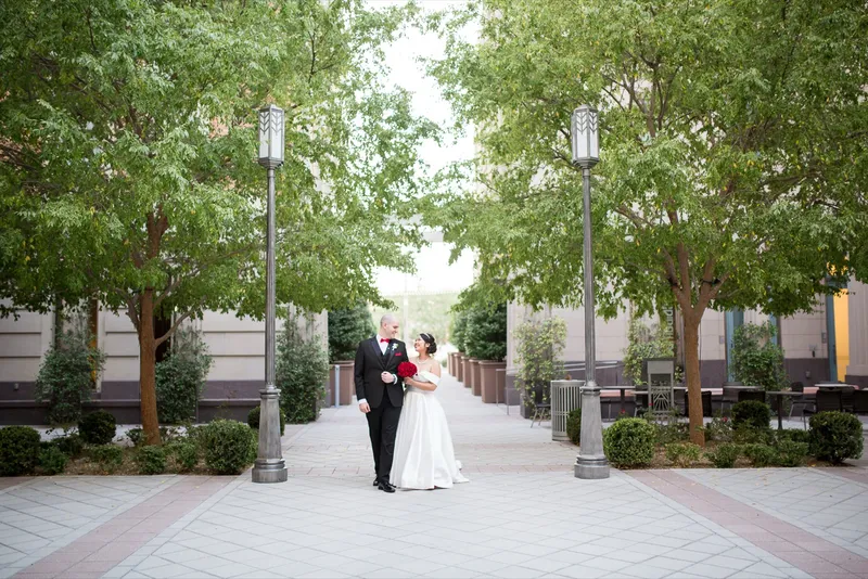 Bride in a satin ballgown and groom in a black tux walking a tree lined plaza between two tall lampposts, patio tables behind them