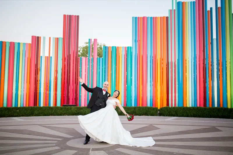 Groom throwing one arm out and laughing as he dips his bride in front of the pipe sculpture, her red bouquet swinging wide