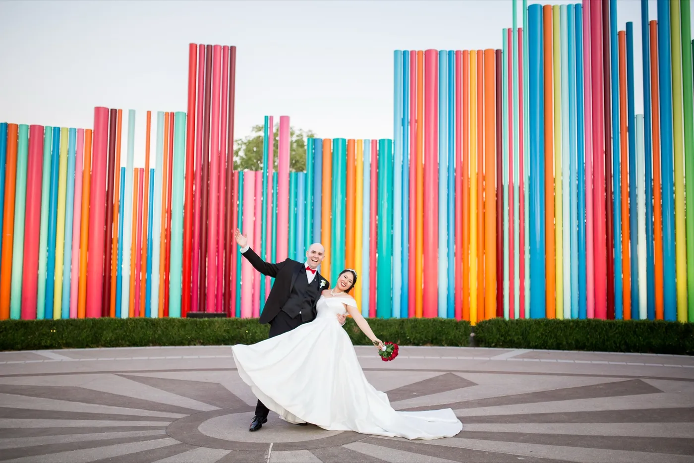 Groom in a black tuxedo throwing an arm out as he dips his bride in a full satin gown in front of a wall of colored pipes