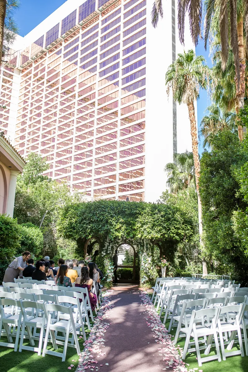 Ceremony aisle strewn with pink and white petals leading to the ivy covered gazebo, white chairs on both sides and the resort's pink glass tower rising behind the trees