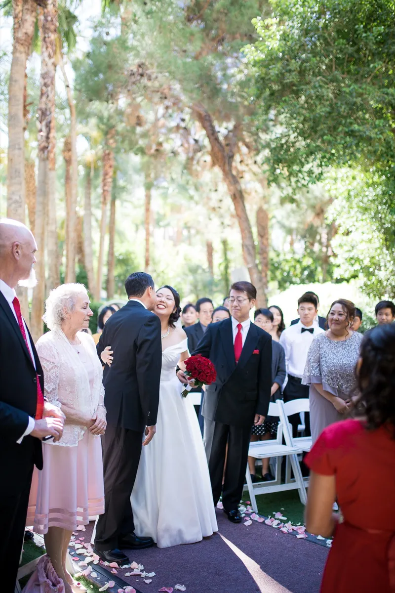 Bride coming up the petal strewn aisle between her parents as seated guests turn to watch