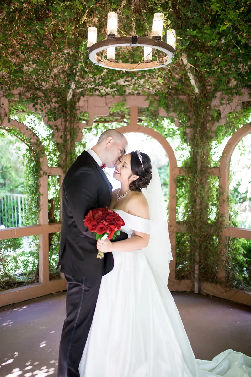 Groom leaning in close to the bride under the gazebo with her red roses at her side