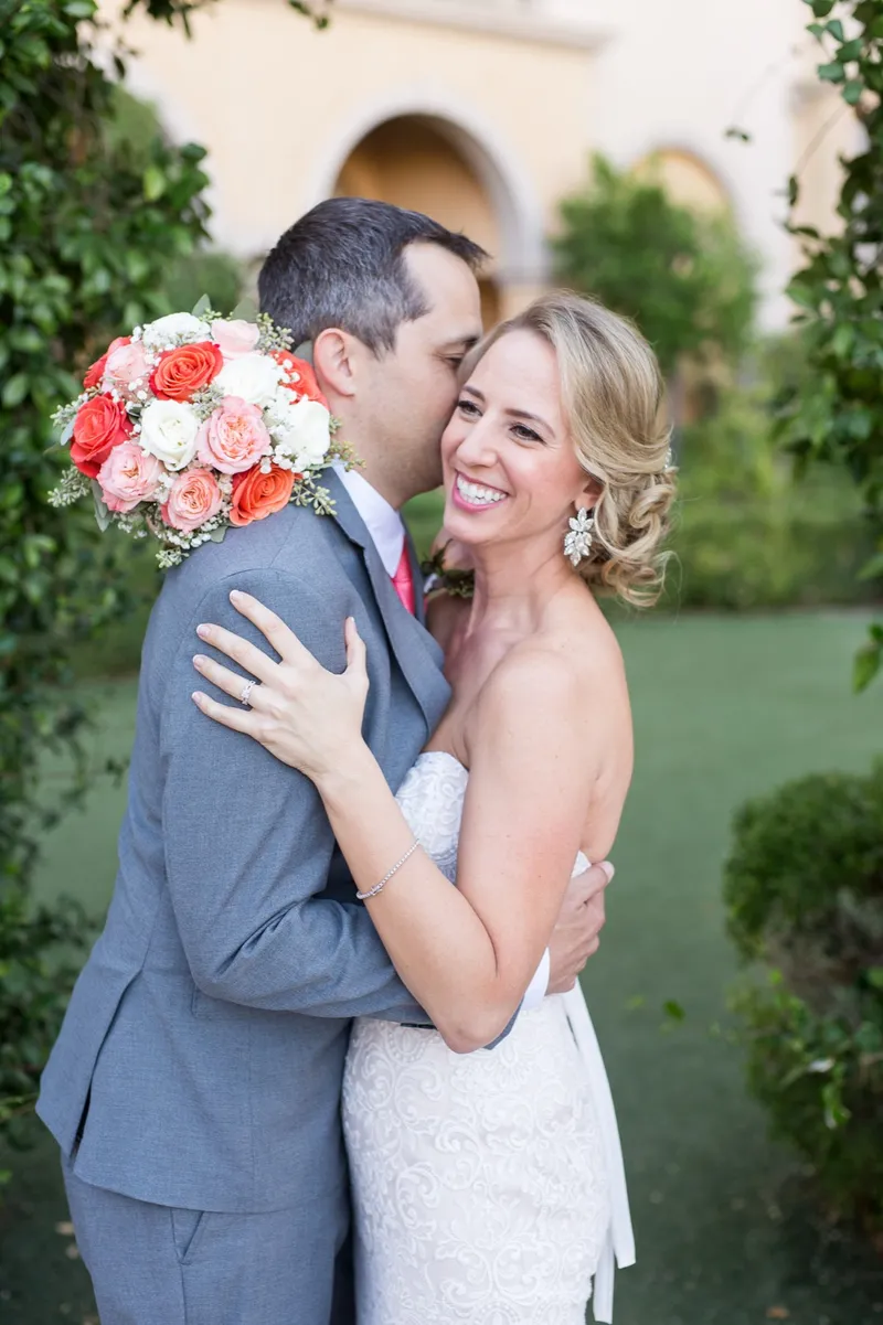 Groom kissing the bride's cheek while she holds a coral and white bouquet