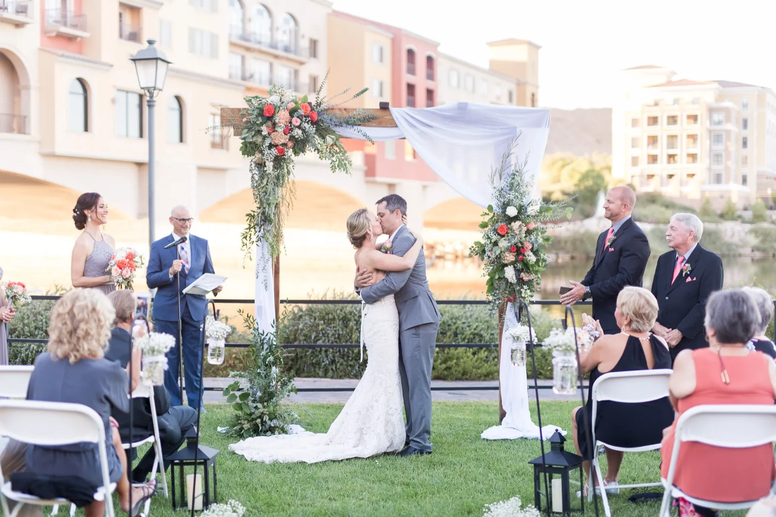 Couple embracing at a floral arch on a lakeside lawn, the village buildings and stone bridge behind them
