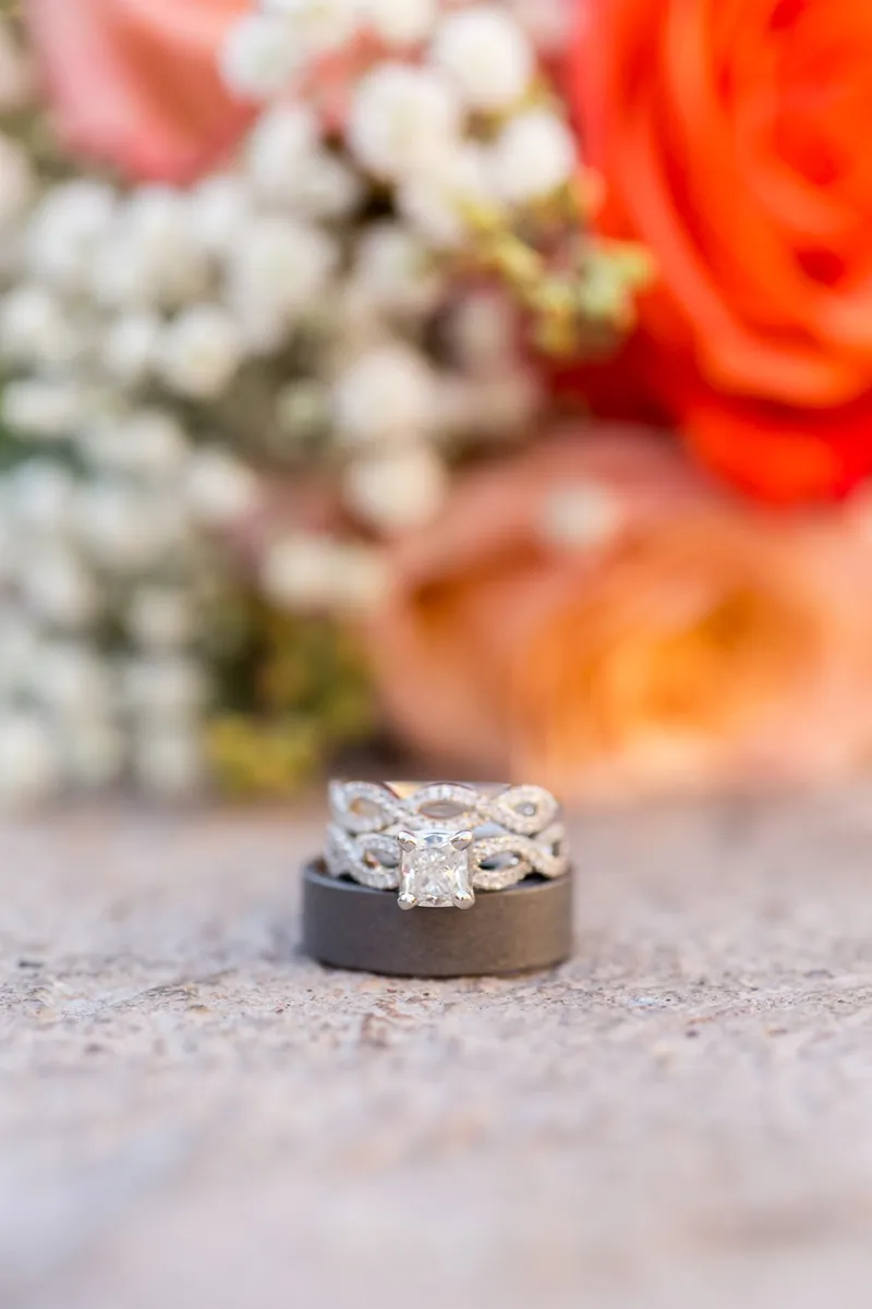 Two rings stacked on a small dark box on stone paving with a coral and white bouquet blurred behind them