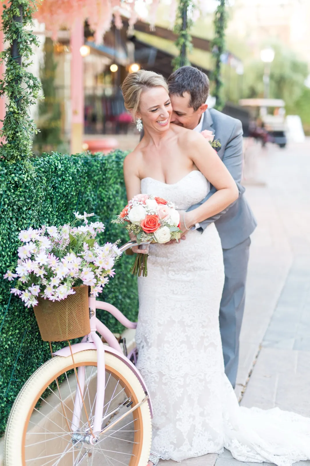 Groom holds his bride from behind beside a pink cruiser bike with a basket of daisies in the village