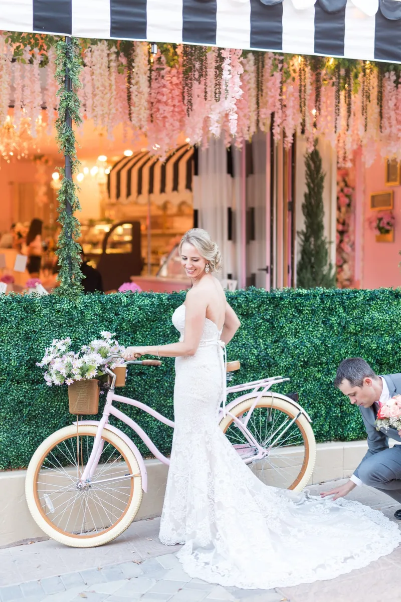 Bride holding the bike upright while the groom crouches to lay out her train outside a village shopfront