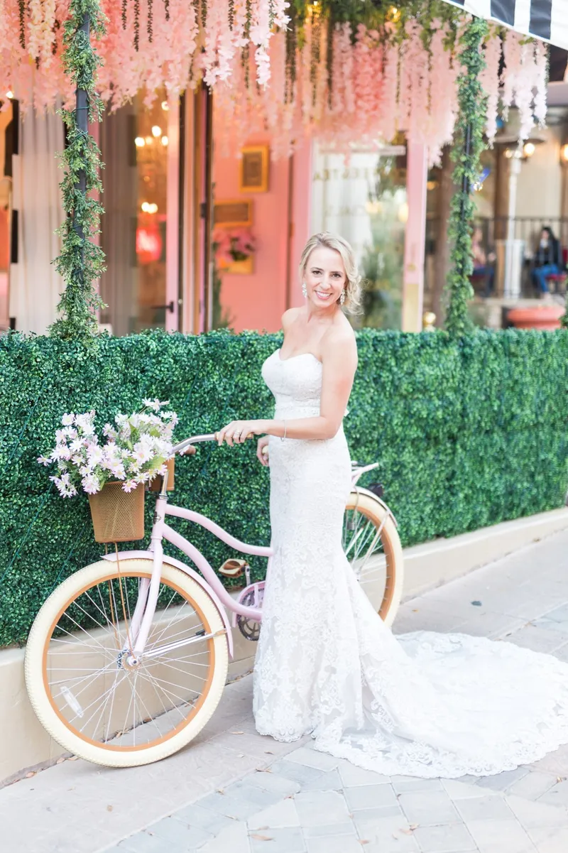 Bride standing with the pink bike in front of a hedge wall on the village street