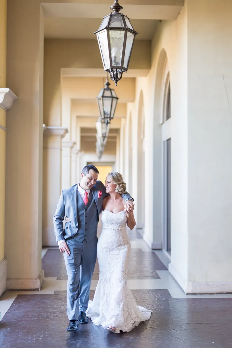 Bride and groom walking the covered walkway together, laughing, her train sweeping the stone floor