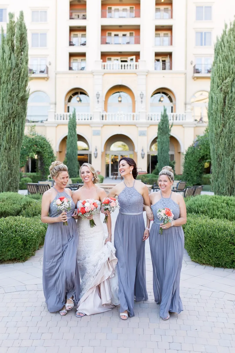 Bride walking through the courtyard with three bridesmaids in dusty blue, the resort arches behind them