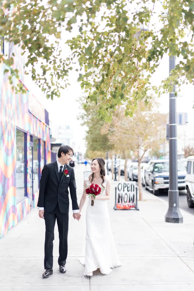 Bride and groom walking a downtown sidewalk under a leafy tree, her red rose bouquet in one hand