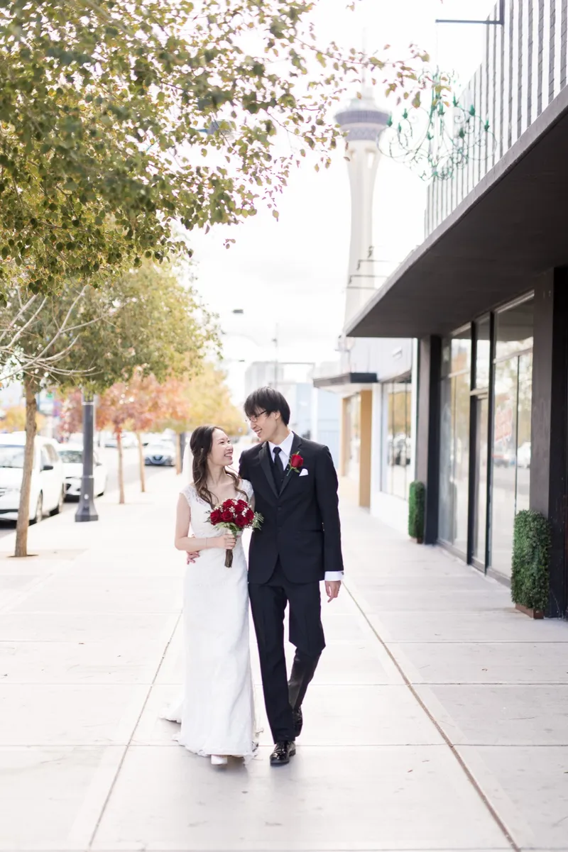 Bride walking a step ahead of her groom past downtown storefronts with her lace gown trailing on the sidewalk