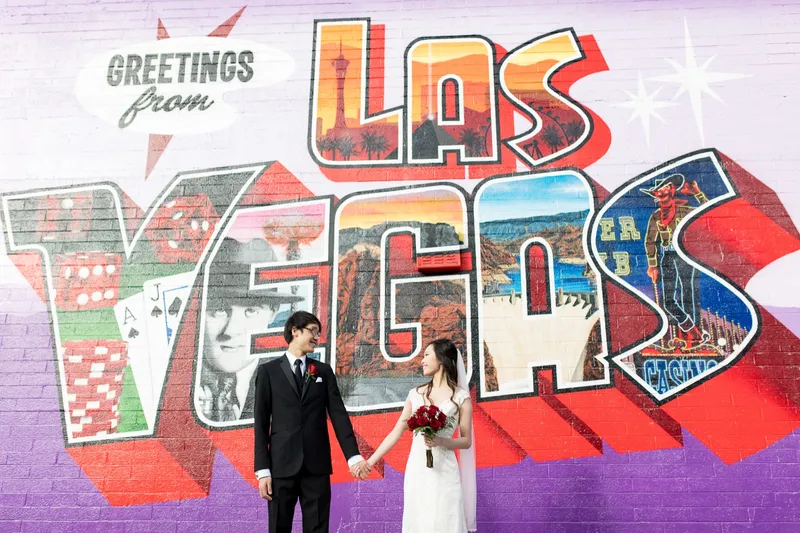 Bride and groom holding hands in front of the large Greetings from Las Vegas postcard mural