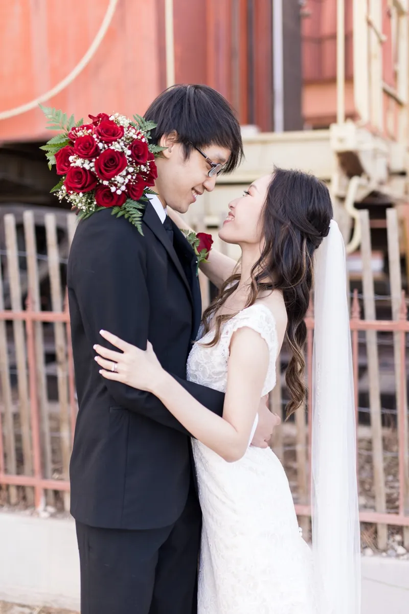 Bride and groom in a close embrace beside a rust red railcar parked behind an iron fence