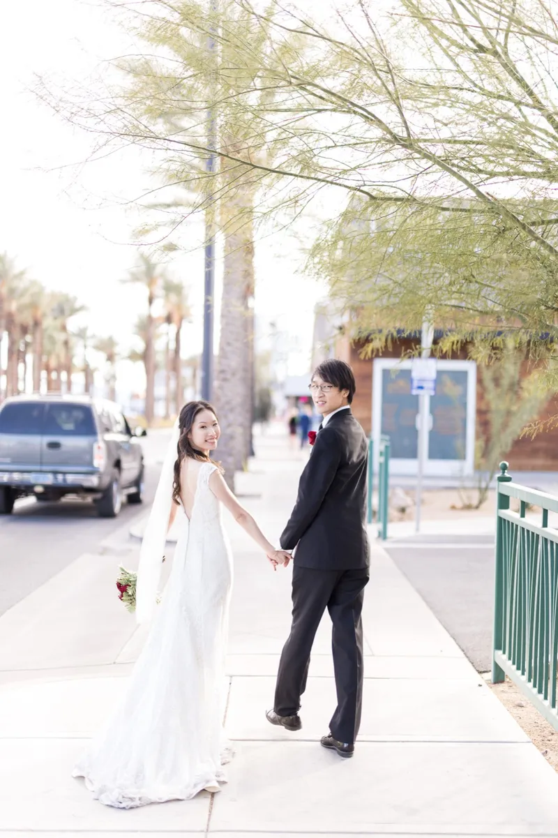 Bride leading her groom by the hand along a sidewalk lined with palm trees, her veil trailing behind her