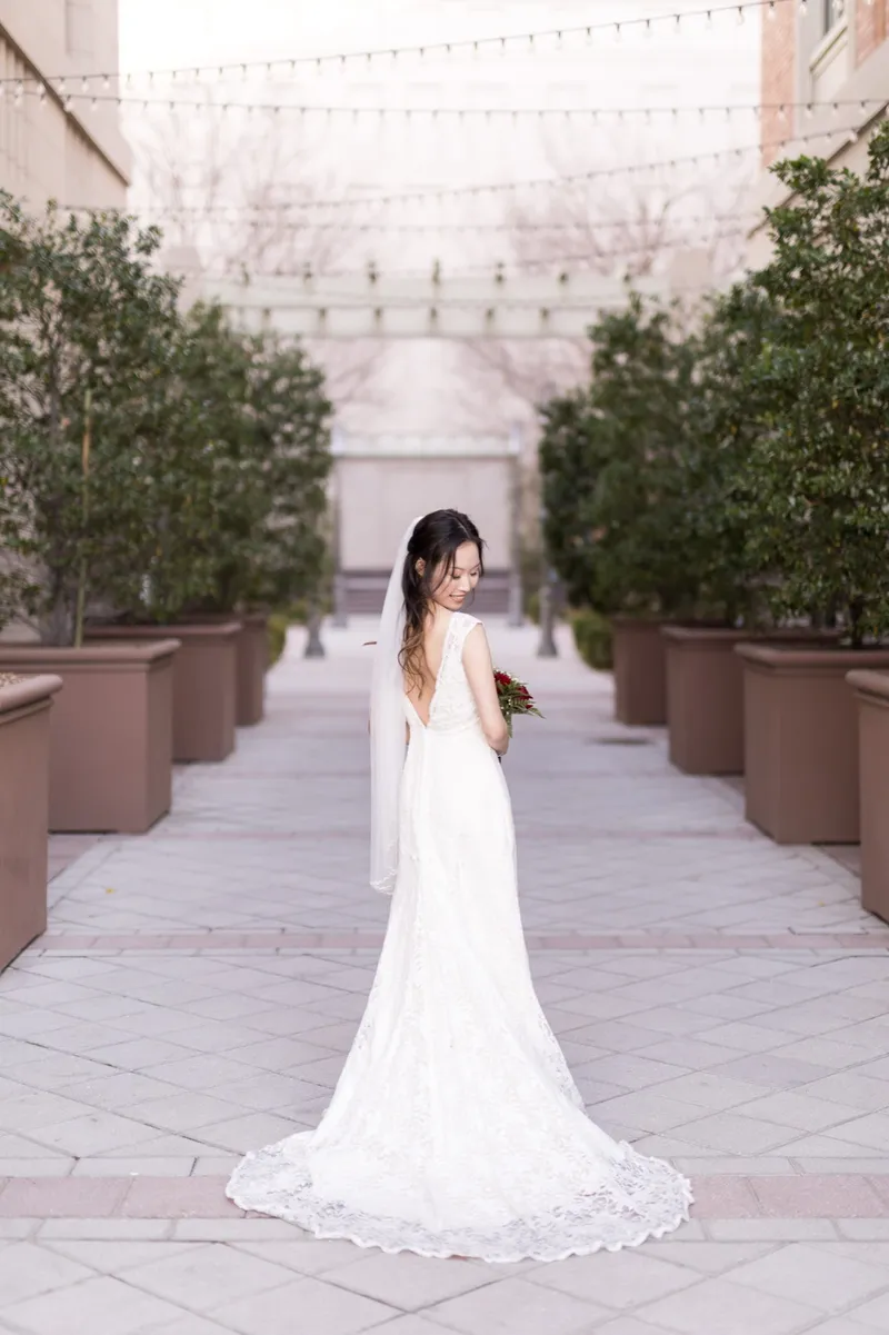 Bride looking back over her bare shoulder in the middle of the planter lined walkway, her lace gown open at the back