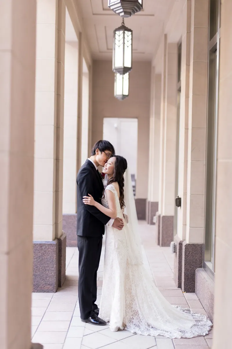 Groom kissing his bride on the forehead in the pale limestone colonnade with art deco lanterns hanging overhead