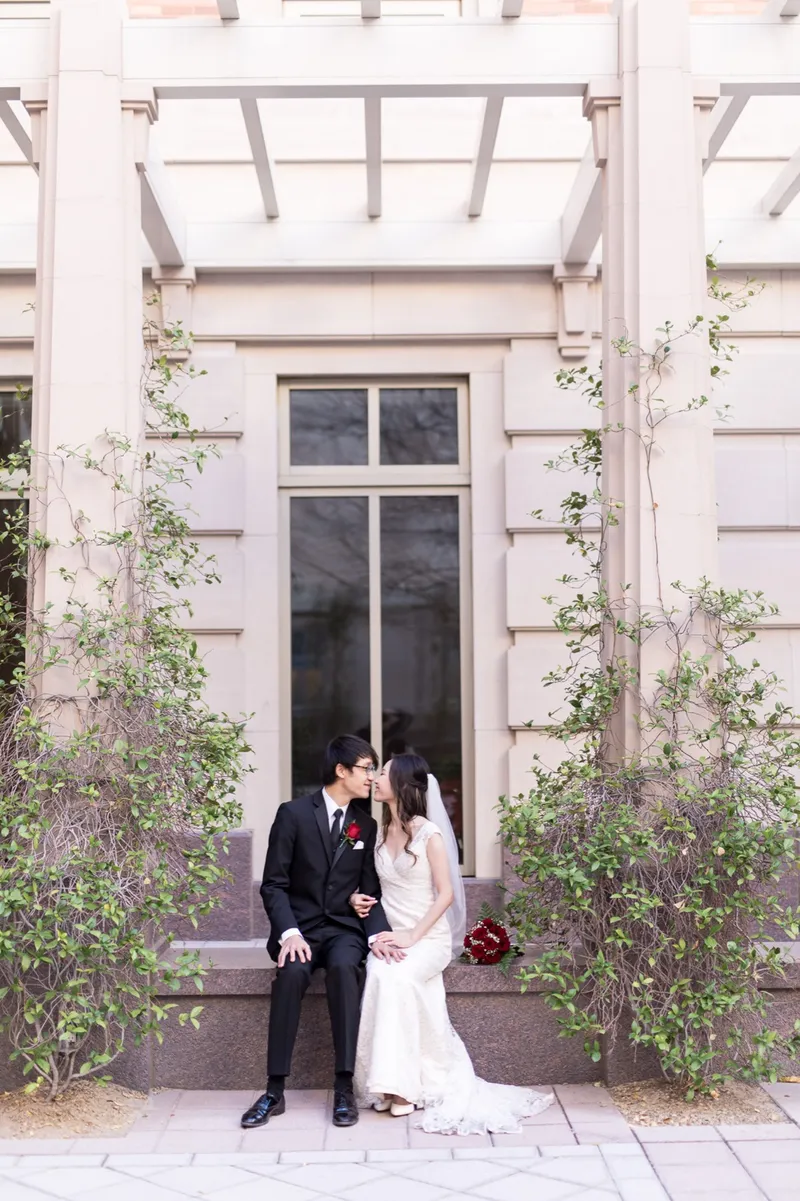 Couple sitting together on the stone ledge under the vine covered wall, her red rose bouquet resting on her lap