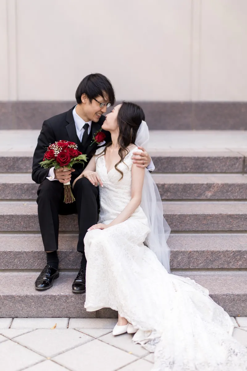 Couple sitting side by side on wide granite steps, her red rose bouquet in her lap and his arm around her