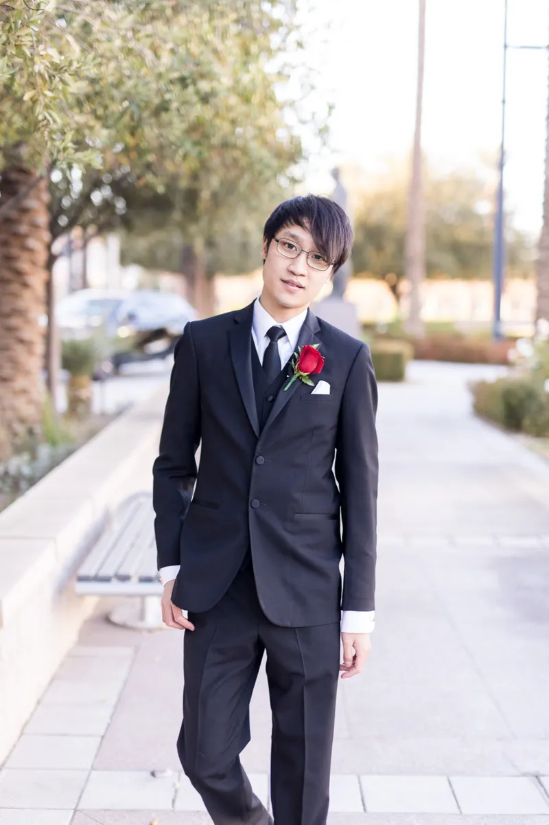 Groom in a black suit with a red rose boutonniere walking the plaza toward the camera, palms and downtown buildings behind him