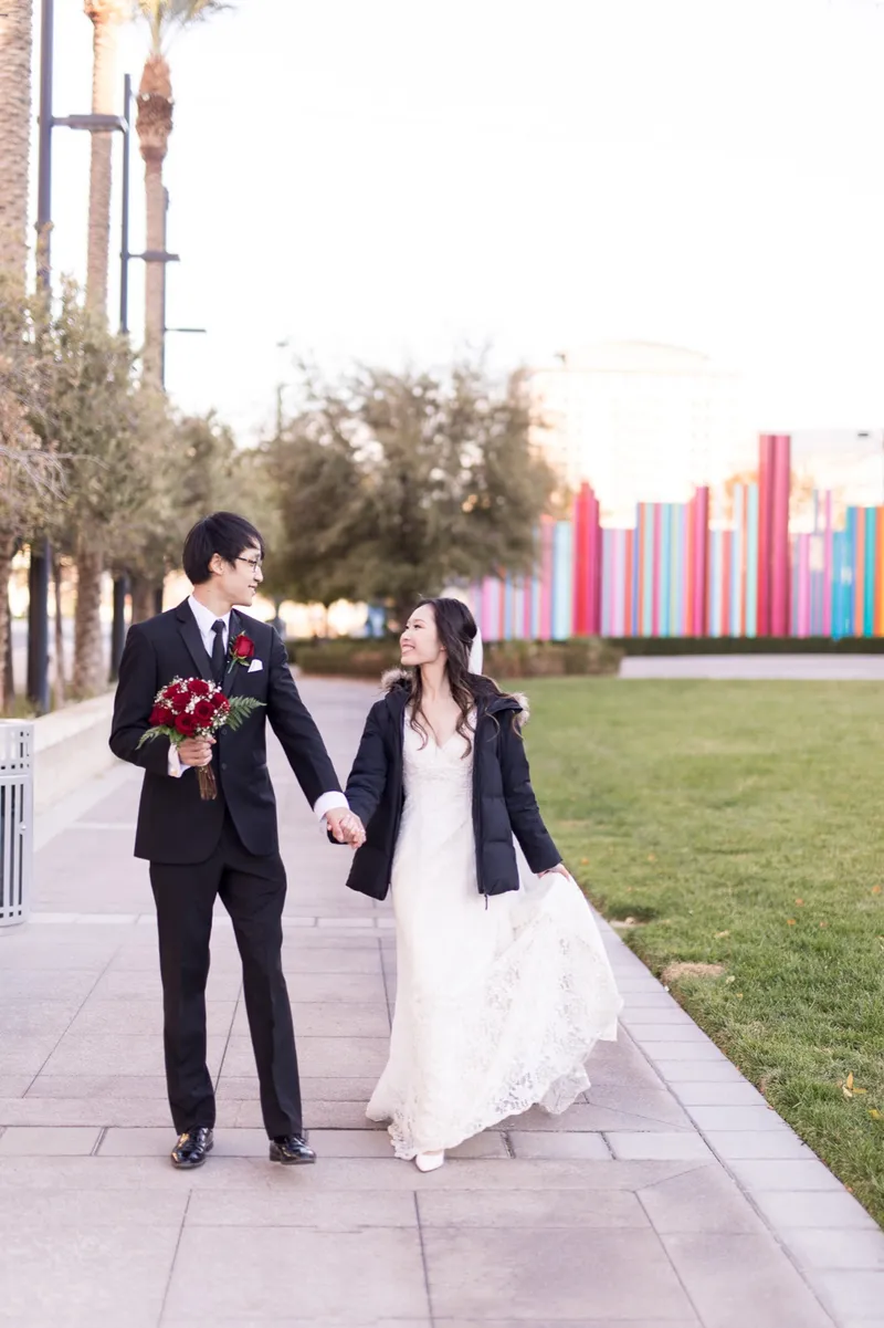 Couple walking a path beside the park lawn with his black jacket over her shoulders and the colored pipes of the sculpture behind them