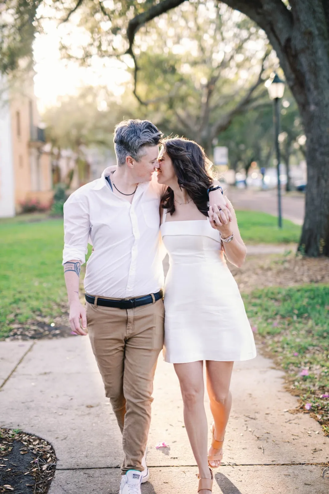 Two women walking a sidewalk under mossy oaks, one in a white button down and chinos, the other in a short white dress