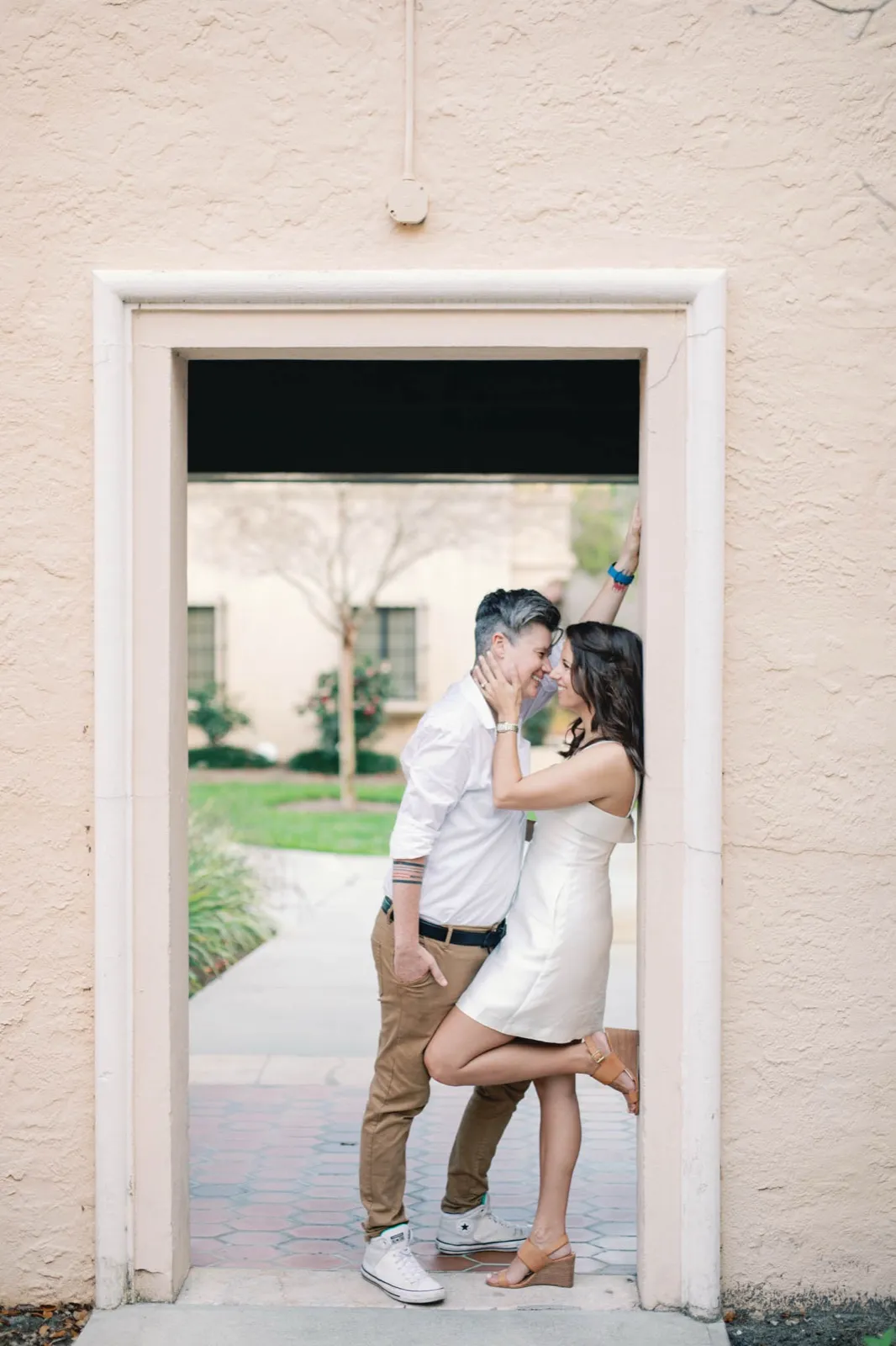 Two women laugh forehead to forehead in a stucco doorway, one in a white shirt, the other lifting a heel