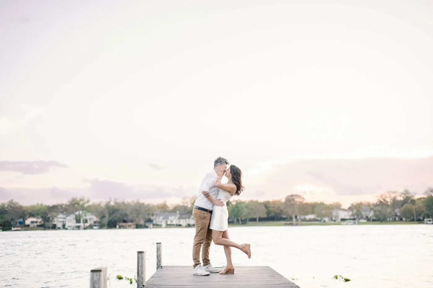 An engaged couple lifted into a joyful kiss on a lakeside dock at sunset
