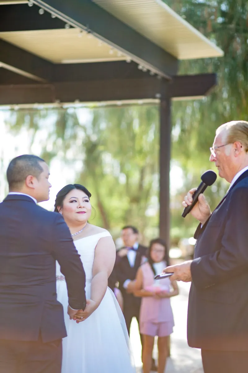 Couple holding hands through their vows under the covered patio with bistro lights strung along the awning above them